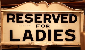 A vintage-style sign with bold black lettering on a white background reads 'Reserved for Ladies.' The sign features classic, ornate edges and subtle detailing, suggesting an old-fashioned or historical context.