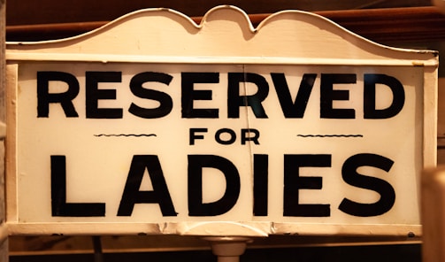 A vintage-style sign with bold black lettering on a white background reads 'Reserved for Ladies.' The sign features classic, ornate edges and subtle detailing, suggesting an old-fashioned or historical context.