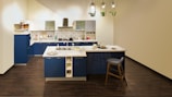A modern kitchen featuring blue cabinets, a white countertop, and stainless steel appliances. There is a central island with a sink, complemented by a high chair. The floor is a dark wood finish, and pendant lights with plants hang from the ceiling.