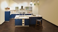 A modern kitchen featuring blue cabinets, a white countertop, and stainless steel appliances. There is a central island with a sink, complemented by a high chair. The floor is a dark wood finish, and pendant lights with plants hang from the ceiling.