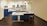 A modern kitchen featuring blue cabinets, a white countertop, and stainless steel appliances. There is a central island with a sink, complemented by a high chair. The floor is a dark wood finish, and pendant lights with plants hang from the ceiling.
