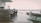 A serene waterfront scene features several boat engines lined up along a dock. The outboard motors bear the Suzuki logo and are positioned near a weathered, rust-colored bollard. In the foreground, a section of a small boat with a rope attached is visible. Across the water, an industrial port can be seen in the distance, with cranes and buildings silhouetted against a hazy sky.