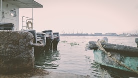 A serene waterfront scene features several boat engines lined up along a dock. The outboard motors bear the Suzuki logo and are positioned near a weathered, rust-colored bollard. In the foreground, a section of a small boat with a rope attached is visible. Across the water, an industrial port can be seen in the distance, with cranes and buildings silhouetted against a hazy sky.