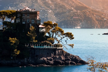 A picturesque castle-like structure is situated on a rocky cliff surrounded by lush greenery and trees. The building overlooks a tranquil blue sea with mountains in the background. The sunlight bathes the scene in a warm glow, enhancing the serene and idyllic atmosphere.