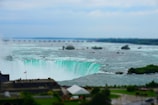 A large waterfall cascades into a river below, surrounded by lush greenery. The powerful water is frothy white with a hint of turquoise as it plunges over the edge. In the foreground, there is a building with a Canadian flag, indicating a location in Canada. The sky above is overcast, adding a serene backdrop to the dramatic falls.