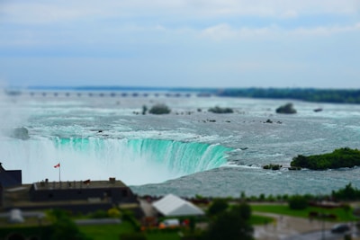 A large waterfall cascades into a river below, surrounded by lush greenery. The powerful water is frothy white with a hint of turquoise as it plunges over the edge. In the foreground, there is a building with a Canadian flag, indicating a location in Canada. The sky above is overcast, adding a serene backdrop to the dramatic falls.