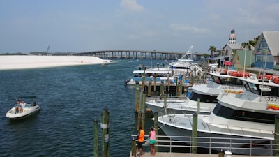 A nautical scene with several boats docked at a marina. A small motorboat with people aboard navigates near the shore. In the background, there is an arched bridge spanning across the water. A lighthouse and some colorful buildings are visible on the right. Two individuals standing on a dock observe the view.