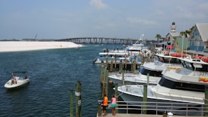 A nautical scene with several boats docked at a marina. A small motorboat with people aboard navigates near the shore. In the background, there is an arched bridge spanning across the water. A lighthouse and some colorful buildings are visible on the right. Two individuals standing on a dock observe the view.