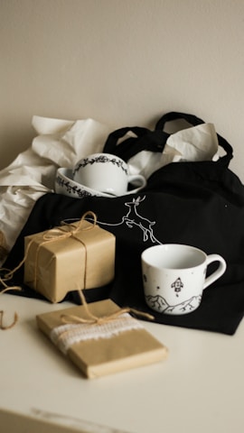 A cozy arrangement featuring two ceramic mugs with black designs, placed on a black cloth bag with a deer illustration. Nearby, there are two brown paper-wrapped gifts tied with twine, and crumpled white tissue paper providing a soft backdrop.