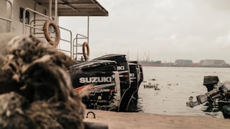A dock with multiple outboard motors attached to a boat, displaying the brand name Suzuki. The scene is set by a waterfront with industrial structures and cranes visible in the distance. The atmosphere appears overcast, with cloudy skies.