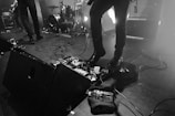 A stage setup during a live performance with musicians, featuring drum set, speakers, and guitar equipment. Cables are scattered across the floor, and a bottle of water is placed near the musical gear.