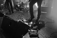 A stage setup during a live performance with musicians, featuring drum set, speakers, and guitar equipment. Cables are scattered across the floor, and a bottle of water is placed near the musical gear.