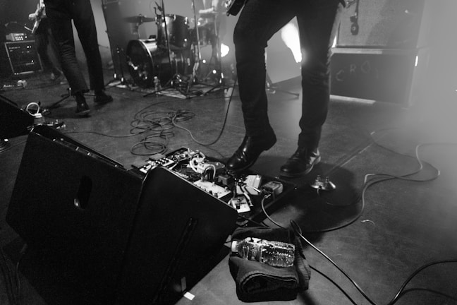 A stage setup during a live performance with musicians, featuring drum set, speakers, and guitar equipment. Cables are scattered across the floor, and a bottle of water is placed near the musical gear.