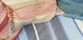 Several textured and colorful fabric cushions are arranged together. The fabrics have different patterns, including stripes, and various weaving styles with fringes.