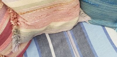 Several textured and colorful fabric cushions are arranged together. The fabrics have different patterns, including stripes, and various weaving styles with fringes.