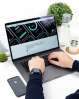 A person is typing on a MacBook Pro laptop surrounded by a smartphone, a green plant, a bottle of VOSS water, and a Starbucks paper cup. The laptop screen displays a UI/UX design with several circular graphic elements and a heading related to an iOS concept. The individual is wearing a smartwatch on their wrist.
