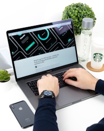 A person is typing on a MacBook Pro laptop surrounded by a smartphone, a green plant, a bottle of VOSS water, and a Starbucks paper cup. The laptop screen displays a UI/UX design with several circular graphic elements and a heading related to an iOS concept. The individual is wearing a smartwatch on their wrist.