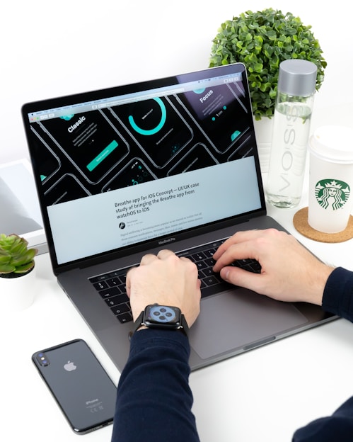 A person is typing on a MacBook Pro laptop surrounded by a smartphone, a green plant, a bottle of VOSS water, and a Starbucks paper cup. The laptop screen displays a UI/UX design with several circular graphic elements and a heading related to an iOS concept. The individual is wearing a smartwatch on their wrist.