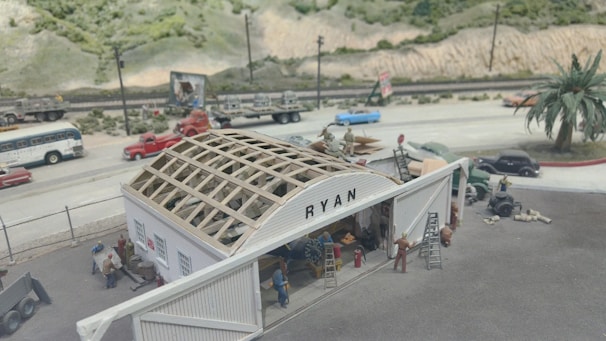 A detailed diorama depicting a construction scene, featuring people working on a building with a partially constructed roof. The structure has the name 'RYAN' on it. There are various vehicles and trees in the background, as well as a road with cars and a bus. The environment includes a mountainous terrain and a railway track along the road.