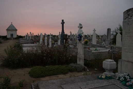 A solemn cemetery scene at dusk with rows of tombstones, some adorned with flowers, and a small chapel-like structure in the distance. Shadows are long, and the sky is a gradient of darkening purple and orange hues as the sun sets.