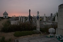 A solemn cemetery scene at dusk with rows of tombstones, some adorned with flowers, and a small chapel-like structure in the distance. Shadows are long, and the sky is a gradient of darkening purple and orange hues as the sun sets.