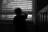 A silhouette of a child looking through a window with blinds partially open, creating contrasting lines of light and shadow. The atmosphere is quiet and introspective, with the child standing next to a piece of furniture.