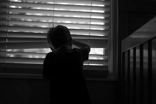 A silhouette of a child looking through a window with blinds partially open, creating contrasting lines of light and shadow. The atmosphere is quiet and introspective, with the child standing next to a piece of furniture.