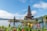 A traditional multi-tiered Balinese temple structure sits on a small island in a tranquil lake surrounded by lush greenery. Brightly colored flowers in the foreground add vibrant contrast to the scene. The sky is mostly clear with a few scattered clouds, creating a peaceful and serene atmosphere.