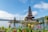A traditional multi-tiered Balinese temple structure sits on a small island in a tranquil lake surrounded by lush greenery. Brightly colored flowers in the foreground add vibrant contrast to the scene. The sky is mostly clear with a few scattered clouds, creating a peaceful and serene atmosphere.