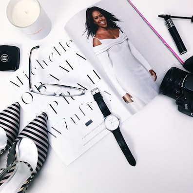 A flat lay arrangement featuring a magazine with a stylish woman in a white outfit, surrounded by a black and white striped pair of shoes, a pair of glasses, a watch, a camera lens, makeup items, and a candle.