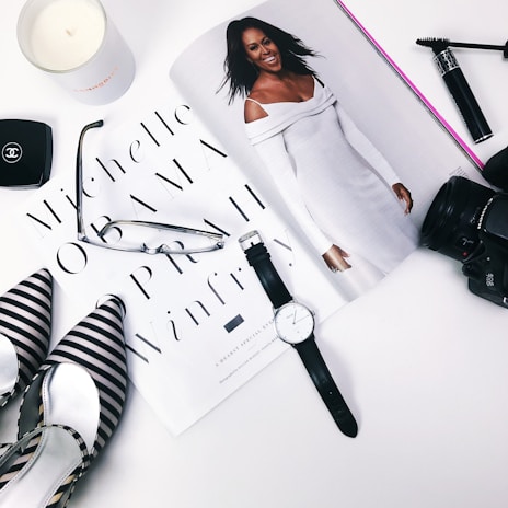 A flat lay arrangement featuring a magazine with a stylish woman in a white outfit, surrounded by a black and white striped pair of shoes, a pair of glasses, a watch, a camera lens, makeup items, and a candle.
