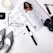 A flat lay arrangement featuring a magazine with a stylish woman in a white outfit, surrounded by a black and white striped pair of shoes, a pair of glasses, a watch, a camera lens, makeup items, and a candle.
