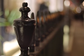 A close-up view of an ornate metal fence post, with a shallow depth of field that creates a blurred, bokeh effect in the background. The focus is on the dark, glossy finish of the post, while the rest of the fence appears as an abstract pattern of soft shapes and colors.
