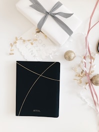 A neatly wrapped gift box sits on a white surface, adorned with a black and white striped ribbon. Beside it is a black notebook with a minimalistic design featuring thin golden lines. Scattered around are decorative elements like gold baubles, red and white ribbons, and gold leaves.