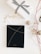 A neatly wrapped gift box sits on a white surface, adorned with a black and white striped ribbon. Beside it is a black notebook with a minimalistic design featuring thin golden lines. Scattered around are decorative elements like gold baubles, red and white ribbons, and gold leaves.
