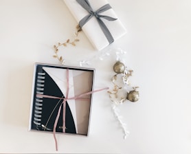 A neatly wrapped notebook with a red and white ribbon is placed in a box. Next to it, a gift wrapped in white paper with a black ribbon is visible. Decorative elements include golden leaf garlands and glittery gold ornaments scattered around, all set against a clean, white background.