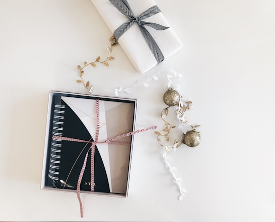 A neatly wrapped notebook with a red and white ribbon is placed in a box. Next to it, a gift wrapped in white paper with a black ribbon is visible. Decorative elements include golden leaf garlands and glittery gold ornaments scattered around, all set against a clean, white background.