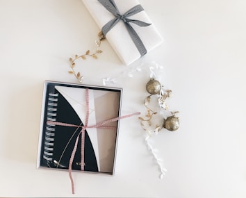 A neatly wrapped notebook with a red and white ribbon is placed in a box. Next to it, a gift wrapped in white paper with a black ribbon is visible. Decorative elements include golden leaf garlands and glittery gold ornaments scattered around, all set against a clean, white background.
