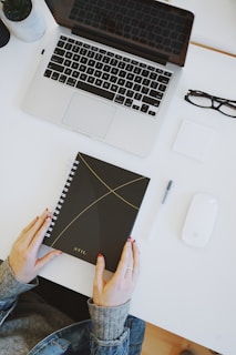 A workspace setup with a laptop, an open spiral-bound notebook with a black cover and minimalist design, a pair of glasses, a pen, and a mouse on a clean white desk. A person wearing a denim jacket is holding the notebook.