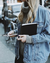 woman standing on sidewalk while holding book and smartphone