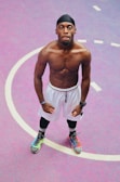A muscular man standing confidently on a vibrant basketball court. He is wearing a black headband, white shorts, leggings, and brightly colored sneakers. The background features a pink and purple surface with white lines marking the court.