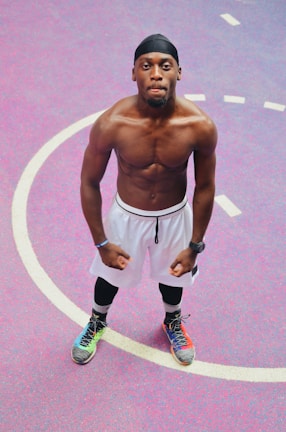 A muscular man standing confidently on a vibrant basketball court. He is wearing a black headband, white shorts, leggings, and brightly colored sneakers. The background features a pink and purple surface with white lines marking the court.