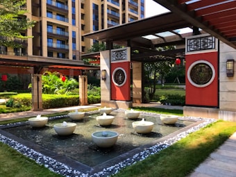 An elegant courtyard features a decorative water fountain surrounded by a tiled edge and landscaped greenery. Red and black architectural accents frame the area, with apartment buildings visible in the background. Traditional Chinese design elements are present, including round decorative panels and hanging red lanterns.