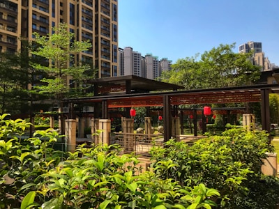 A beautifully landscaped garden area surrounded by modern high-rise buildings. The scene features lush green foliage, a wooden pergola structure adorned with red lanterns, and bright sunlight casting shadows. The sky is clear and blue, enhancing the vibrant colors of the garden.