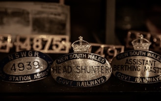 A collection of vintage metallic nameplates featuring inscriptions such as 'New South Wales Head Shunter' and 'Assistant Berthing Master'. The plates are oval-shaped with crowns depicted on top, suggestive of a historical or official context, possibly related to railway positions. The lighting is dim, highlighting the reflective surfaces of the metal.