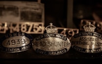 A collection of vintage metallic nameplates featuring inscriptions such as 'New South Wales Head Shunter' and 'Assistant Berthing Master'. The plates are oval-shaped with crowns depicted on top, suggestive of a historical or official context, possibly related to railway positions. The lighting is dim, highlighting the reflective surfaces of the metal.