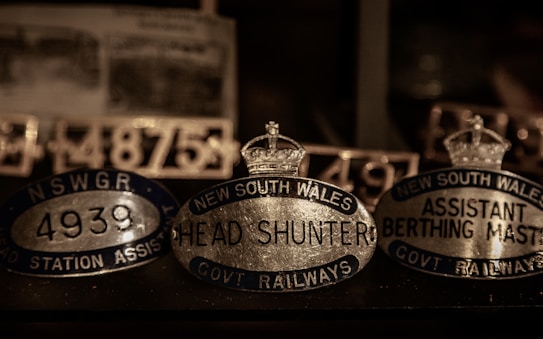 A collection of vintage metallic nameplates featuring inscriptions such as 'New South Wales Head Shunter' and 'Assistant Berthing Master'. The plates are oval-shaped with crowns depicted on top, suggestive of a historical or official context, possibly related to railway positions. The lighting is dim, highlighting the reflective surfaces of the metal.