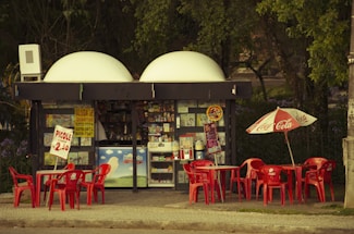 A small kiosk surrounded by trees featuring a variety of signs and advertisements is depicted. Bright red tables and chairs are positioned outside, some beneath a Coca Cola-branded umbrella. There are colorful posters and products displayed inside the kiosk.
