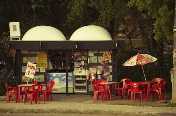 A small kiosk surrounded by trees featuring a variety of signs and advertisements is depicted. Bright red tables and chairs are positioned outside, some beneath a Coca Cola-branded umbrella. There are colorful posters and products displayed inside the kiosk.