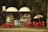 A small kiosk surrounded by trees featuring a variety of signs and advertisements is depicted. Bright red tables and chairs are positioned outside, some beneath a Coca Cola-branded umbrella. There are colorful posters and products displayed inside the kiosk.